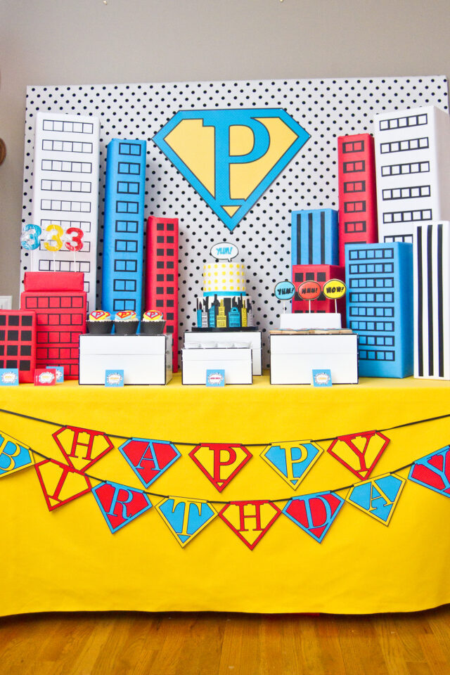 Vintage pop art Super Hero birthday party dessert table with comic book decorations, colorful city skyline backdrop, superhero cake, cupcakes, and candy bar.