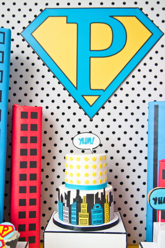 Vintage pop art Super Hero birthday party dessert table with comic book decorations, colorful city skyline backdrop, superhero cake, cupcakes, and candy bar.