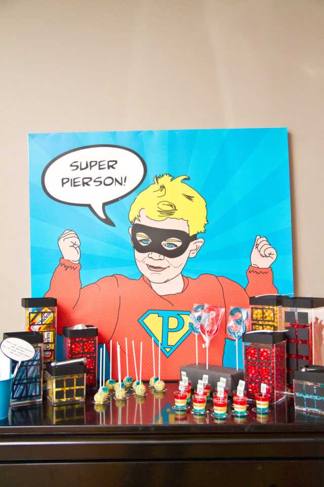 Vintage pop art Super Hero birthday party dessert table with comic book decorations, colorful city skyline backdrop, superhero cake, cupcakes, and candy bar.
