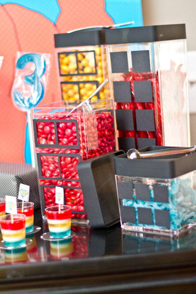 Vintage pop art Super Hero birthday party dessert table with comic book decorations, colorful city skyline backdrop, superhero cake, cupcakes, and candy bar.