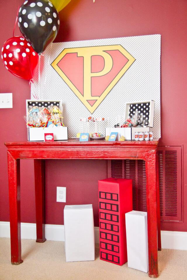 Vintage pop art Super Hero birthday party dessert table with comic book decorations, colorful city skyline backdrop, superhero cake, cupcakes, and candy bar.