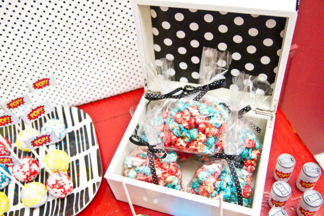 Vintage pop art Super Hero birthday party dessert table with comic book decorations, colorful city skyline backdrop, superhero cake, cupcakes, and candy bar.