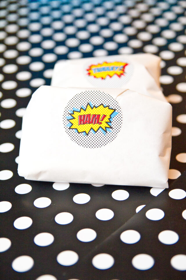 Vintage pop art Super Hero birthday party dessert table with comic book decorations, colorful city skyline backdrop, superhero cake, cupcakes, and candy bar.