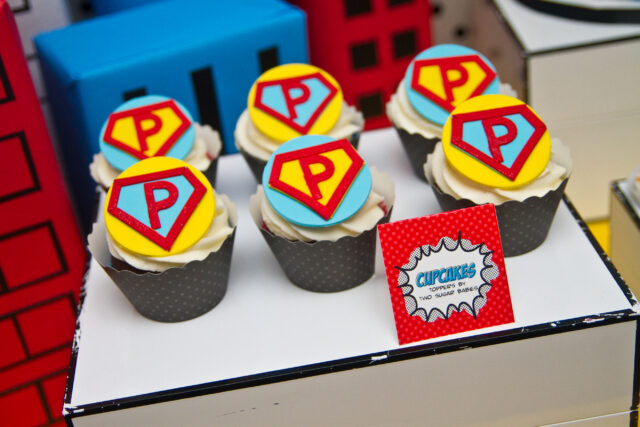 Vintage pop art Super Hero birthday party dessert table with comic book decorations, colorful city skyline backdrop, superhero cake, cupcakes, and candy bar.